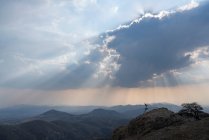 Ein Mann läuft unter wolkenverhangenem Himmel mit Sonnenstrahlen auf einem Felsen bergab — Stockfoto