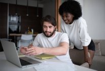 Chico barbudo alegre mostrando datos en el ordenador portátil a la mujer afroamericana mientras trabajan en el proyecto remoto en casa juntos - foto de stock