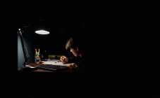 Adolescent garçon dessin à un bureau dans un sombre chambre par lampe lumière. — Photo de stock