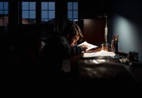 Мальчик-подросток рисует за столом в темной комнате при свете лампы. — стоковое фото