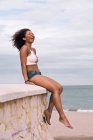 Молода чорна жінка сміється, сидячи на кам'яній стіні біля моря — стокове фото