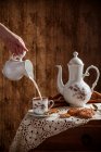 Рука наливая молоко в кофе подается на винтажную посуду с домашним печеньем на деревенском столе с деревянным фоном. — стоковое фото