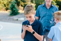 Adolescente menino olha para seu telefone inteligente enquanto fora com família — Fotografia de Stock