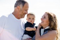 Abuelos sonriendo y sosteniendo nieto en la playa - foto de stock