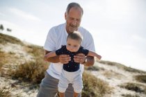 Abuelo jugando y sosteniendo nieto mientras está de pie en la playa - foto de stock