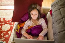 Una niña pequeña se sienta en el sofá en una pila de mantas trabajando en la computadora - foto de stock