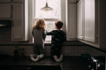 Frère et sœur regardant par la fenêtre de la cuisine — Photo de stock