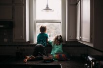 Menino e menina olhando para fora janela da cozinha no dia chuvoso — Fotografia de Stock