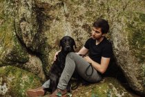 Young man with a black labrador retriever sitting on a mossy rock — Stock Photo