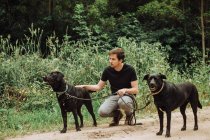 Millennial homem sentado com seus dois cães negros em alerta na floresta — Fotografia de Stock