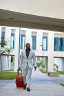Афроамериканец в белом костюме и красном чемодане. Бизнесмен. Бизнесмен путешествует по делам. Путешественник. — стоковое фото