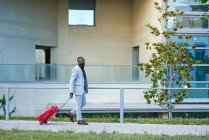 Афроамериканец в белом костюме и красном чемодане. Бизнесмен. Бизнесмен путешествует по делам. Путешественник. — стоковое фото