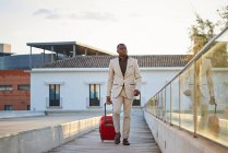 Homme afro-américain en costume élégant et une valise rouge marchant sur une passerelle en bois au coucher du soleil. Homme d'affaires voyageant. — Photo de stock