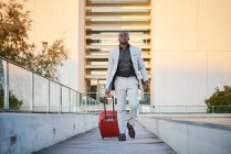 African-American man in a stylish suit and a red suitcase walking down a wooden walkway at sunset. Business man traveling. — Stock Photo