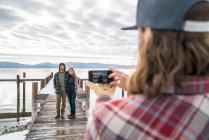 Friend takes a cellphone picture for a couple standing at the end of a pier — Stock Photo