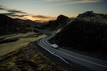Veículo moderno dirigindo na estrada de asfalto sinuosa através de terreno montanhoso pitoresco durante o pôr do sol no campo — Fotografia de Stock