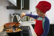Девушка в шляпе красного шеф-повара жарит мясо на сковородке дома — стоковое фото