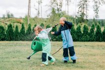 Brother and sister playing in the rain with a hose and watering can — Stock Photo