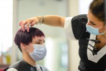 Estilista de cabello femenino en el trabajo con mascarilla mientras peina a una chica joven - foto de stock