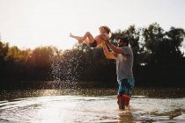 Отец держит маленького ребенка в воздухе у озера, брызгая водой — стоковое фото