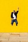 Молодий чоловік з сонцезахисними окулярами стрибає перед жовтою стіною . — стокове фото