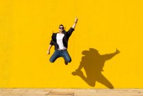 Jeune homme avec des lunettes de soleil sautant devant un mur jaune. — Photo de stock