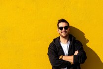 Jeune homme barbu debout contre un mur jaune avec les bras croisés — Photo de stock