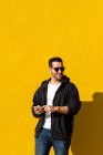 Homem barbudo com óculos de sol em pé contra a parede amarela, usando telefone — Fotografia de Stock