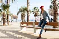 Улыбающаяся женщина в шерстяной шляпе, опирающаяся на скамейку на набережной, отворачивающаяся — стоковое фото