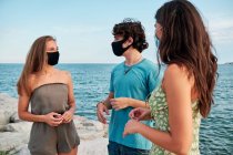Dos chicas jóvenes y un hombre hablando con distancia social y mascarilla en la playa - foto de stock