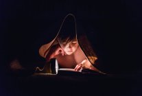 Giovane ragazzo che legge un libro sotto una coperta con una torcia elettrica. — Foto stock