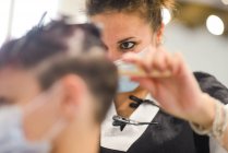 Estilista de cabello femenino en el trabajo con mascarilla mientras peina a una chica joven - foto de stock