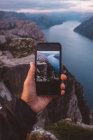 Mão segurando smartphone com imagem de cena no fundo sobre ele em fiordes noruegueses. — Fotografia de Stock
