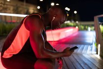 Вид збоку чорного бігуна з навушниками, що використовують телефон під час тренувань вночі — стокове фото
