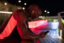 Atleta étnico ouvindo música e navegando telefone durante o intervalo no treino noturno — Fotografia de Stock