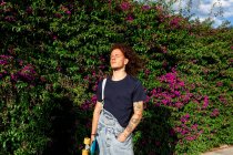 Rotgelockte Haare tätowieren Männer mit Skateboard gegen Pflanzenwand — Stockfoto