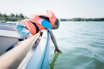Мальчик, окунувший руки в воду летом на лодке — стоковое фото