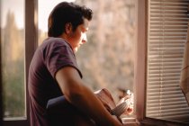 Young man playing guitar while looking out of the window while standing on the balcony at home during self isolation — Stock Photo