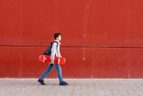 Мальчик держит красный скейтборд, когда идет против красной стены — стоковое фото