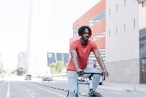 Vista laterale del ciclista afroamericano di sesso maschile in sella alla bicicletta sul terrapieno soleggiato — Foto stock