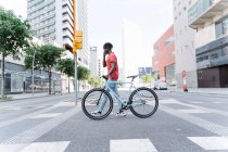 Side view of young ethnic male crossing street with bike and talking on smartphone — Stock Photo
