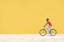 Вид сбоку этнических мужчин на велосипеде на тротуаре возле желтой стены в городе — стоковое фото