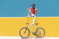 Молодой черный парень сидит на желтой стене возле велосипеда против голубого неба в городе — стоковое фото
