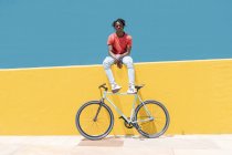 Молодий чорний хлопець сидить на жовтій стіні біля велосипеда проти блакитного неба в місті — стокове фото