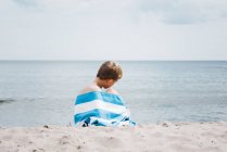 Jeune garçon assis enveloppé dans une serviette rayée seul sur la plage — Photo de stock