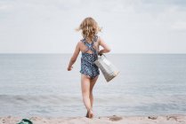 Young girl holding a beach bag strutting towards the sea — Stock Photo