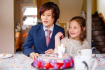 Two smiling children sit at a table in front of lit birthday cake — Stock Photo