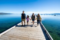 Una familia sonríe y toma las manos caminando por un muelle en un día soleado en South Lake Tahoe, California. - foto de stock
