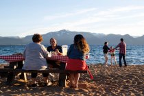 Uma mãe e avós desfrutam de uma conversa durante o jantar enquanto o pai e suas filhas brincam na areia durante o pôr do sol em Nevada Beach, em Lake Tahoe, Nevada. — Fotografia de Stock