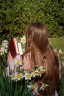 Backview of brown-haired girl reading a book sitting among flowers — Stock Photo